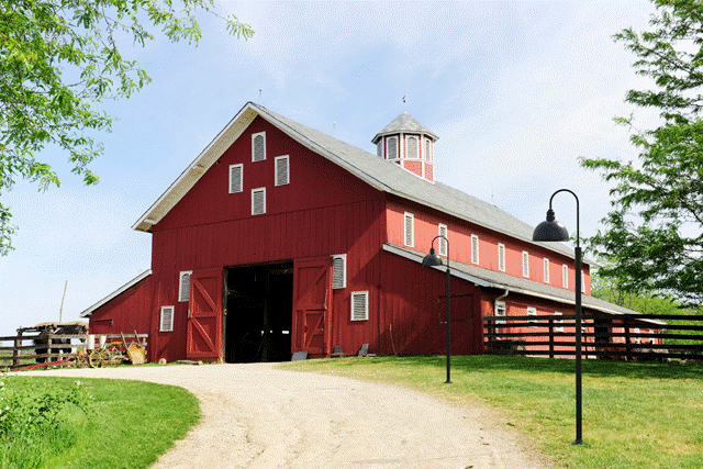 Red barn with gooseneck outdoor lights on a sunny day, surrounded by green trees and a dirt path.