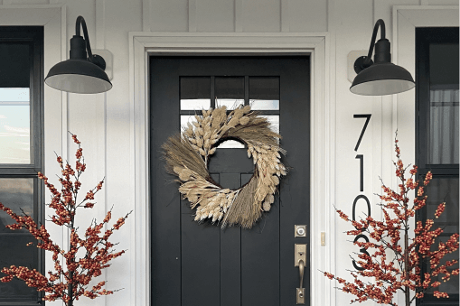 Farmhouse entrance with fall wreath and dual gooseneck barn lights against a white exterior.