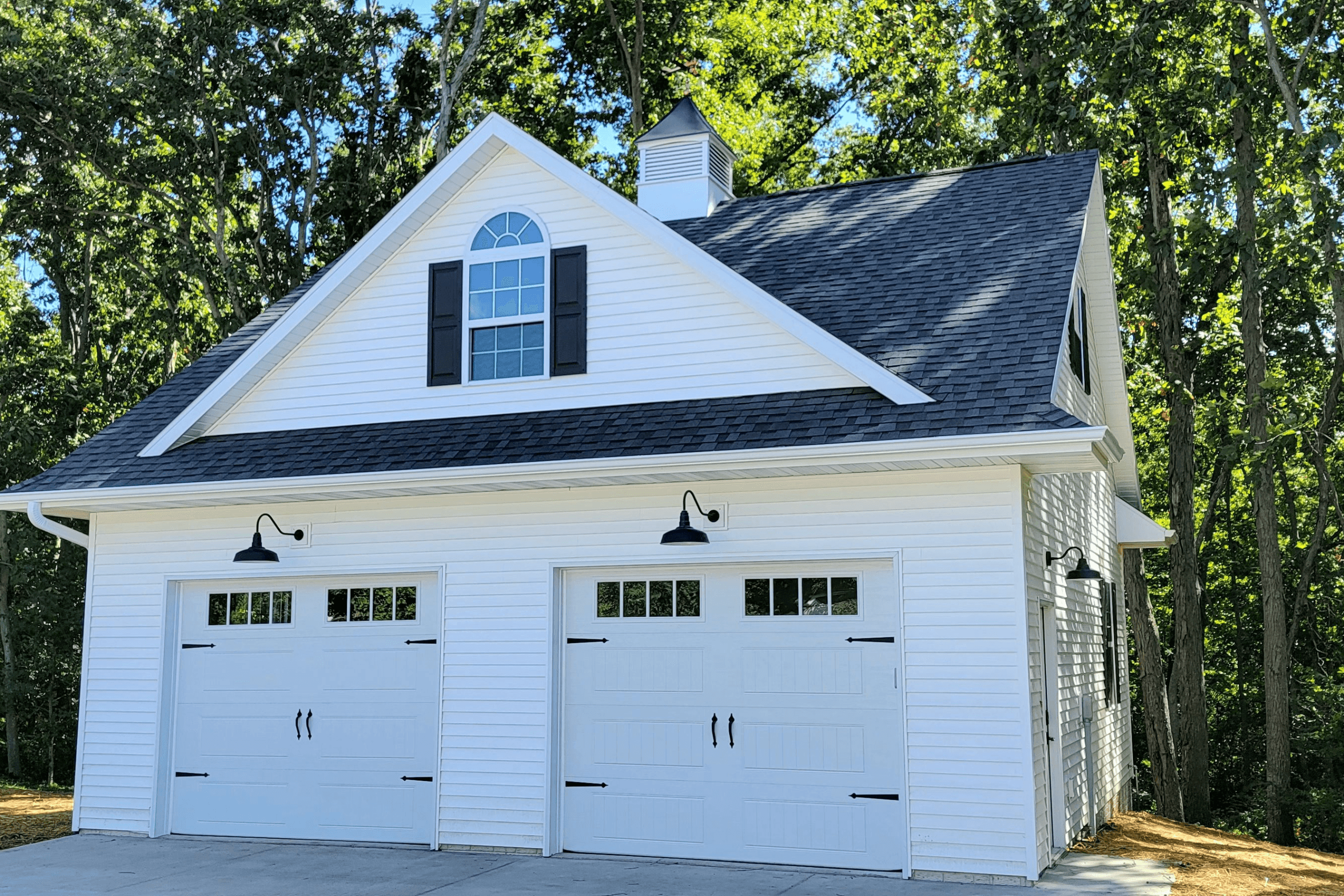 White garage with black gooseneck barn lights, surrounded by trees on a sunny day.