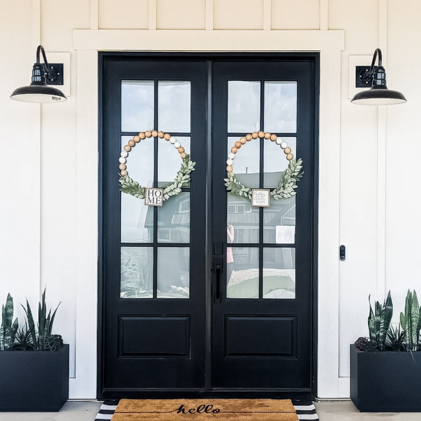 Black double doors with decorative wreaths, flanked by gooseneck outdoor lights and plants.