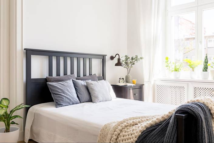 Cozy bedroom with a black bed frame, white linens, and a wall-mounted light beside a sunlit window.