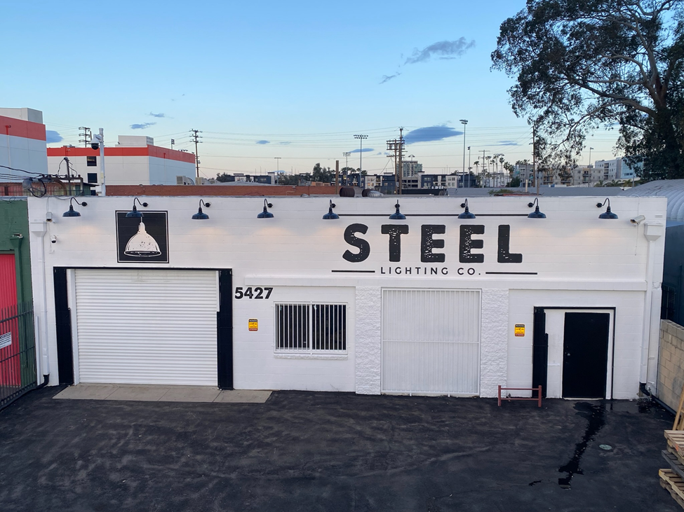 Steel Lighting Co. building exterior with black gooseneck lights mounted on a white facade, cityscape in background.