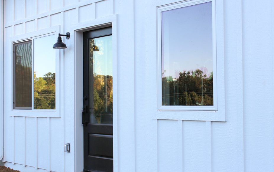 White exterior wall with a black gooseneck barn light beside a door and two windows.